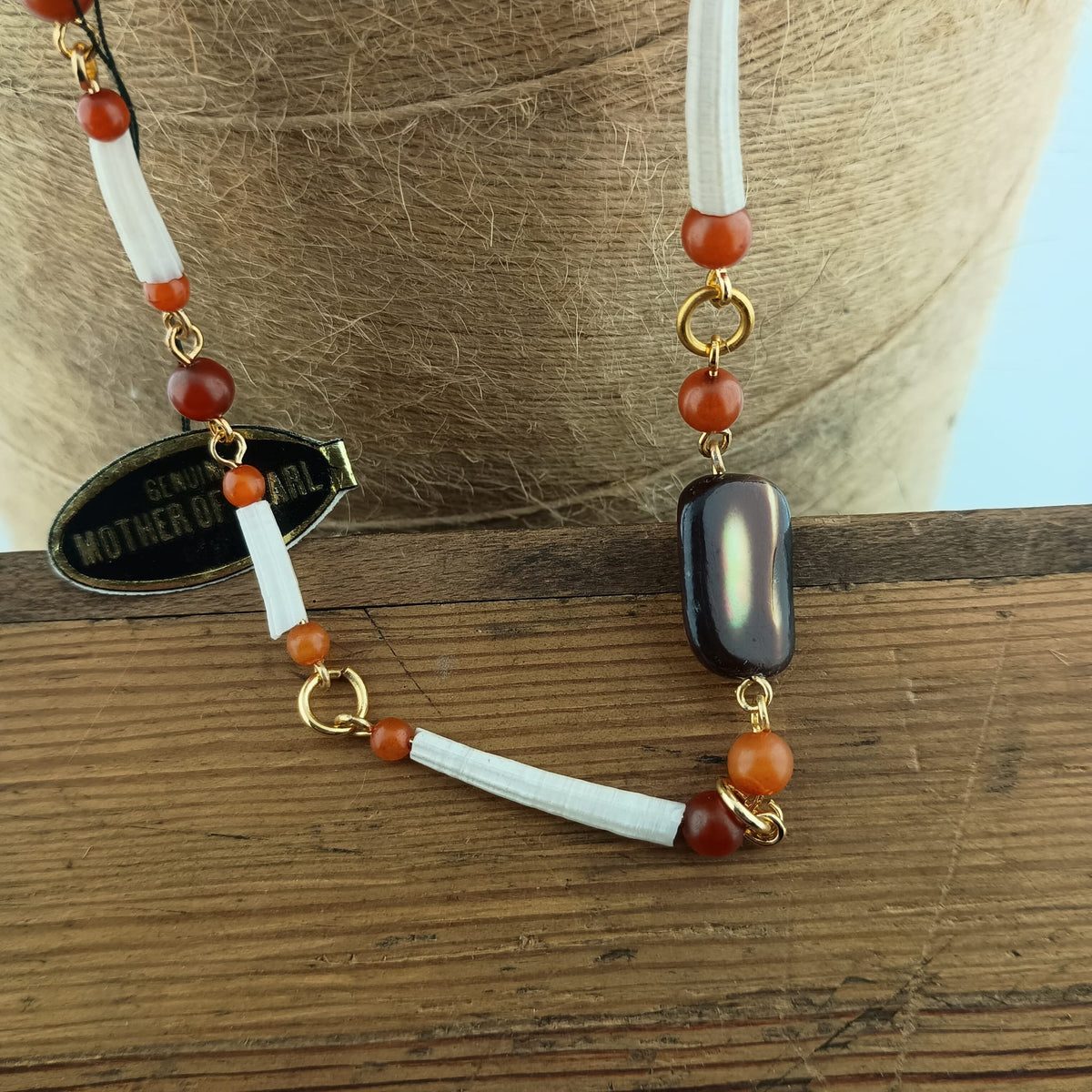 A beaded necklace displayed on a wooden surface, featuring white textured beads, orange-red marbled beads, a large polished black stone centerpiece, and gold-tone metal rings and connectors. A small oval tag reads 'GENUINE MOTHER OF ARIL'.