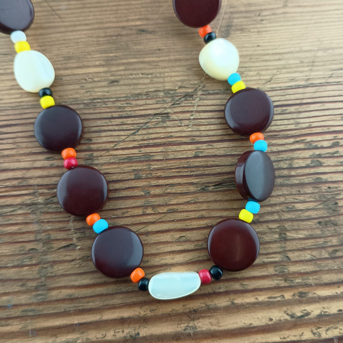 A beaded necklace with large dark reddish-brown round beads, white oval beads, and small multicolored spacer beads, displayed on a wooden surface.