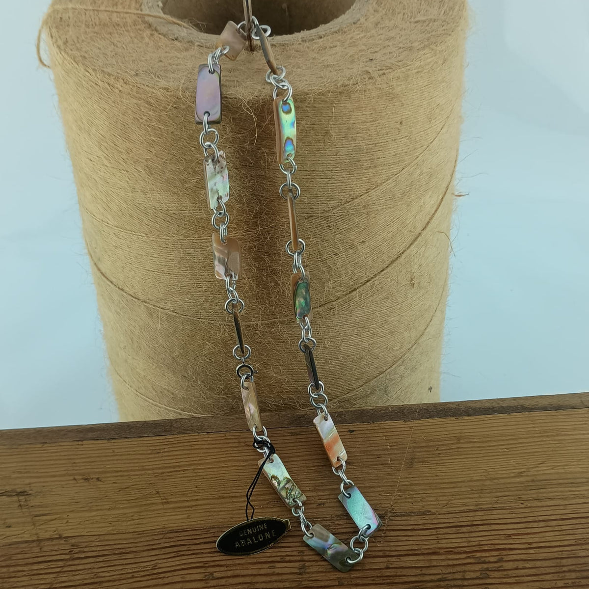 A handmade beaded necklace with silver-toned chain links and natural abalone shell beads in various colors, hanging from a spool of twine on a wooden surface.