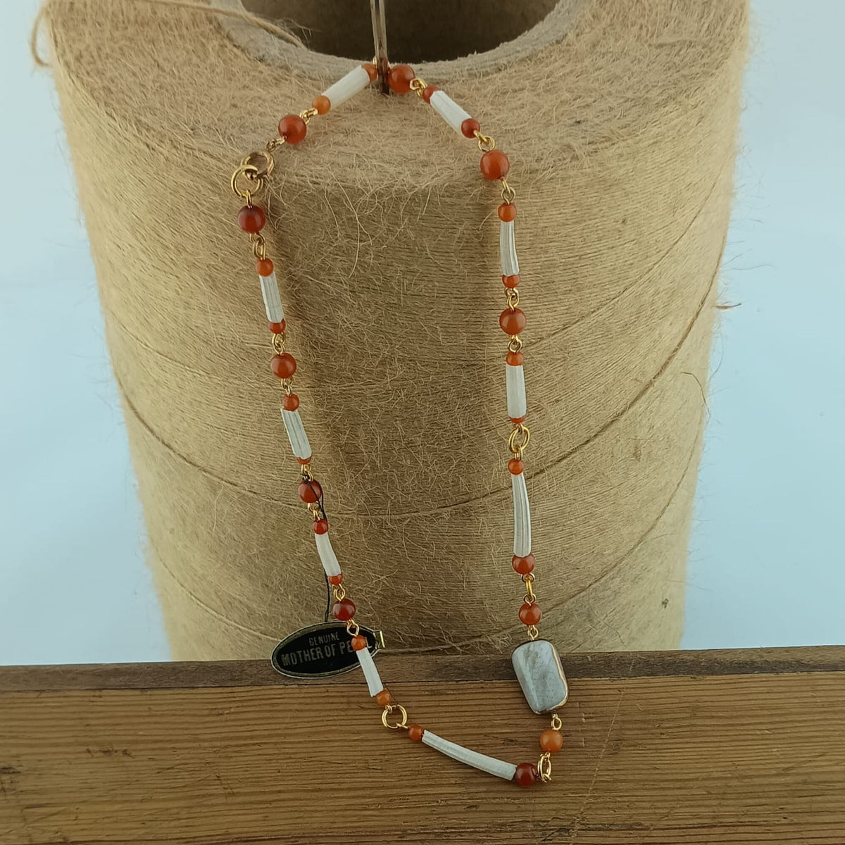 A beaded necklace with white cylindrical shell beads, orange-red round beads, and gold-toned accents, draped over a spool of tan thread on a wooden surface.