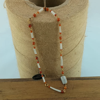 A beaded necklace with white cylindrical shell beads, orange-red round beads, and gold-toned accents, draped over a spool of tan thread on a wooden surface.