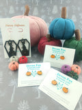 Pumpkin-shaped earrings and cards on a textured surface with decorative pumpkins.