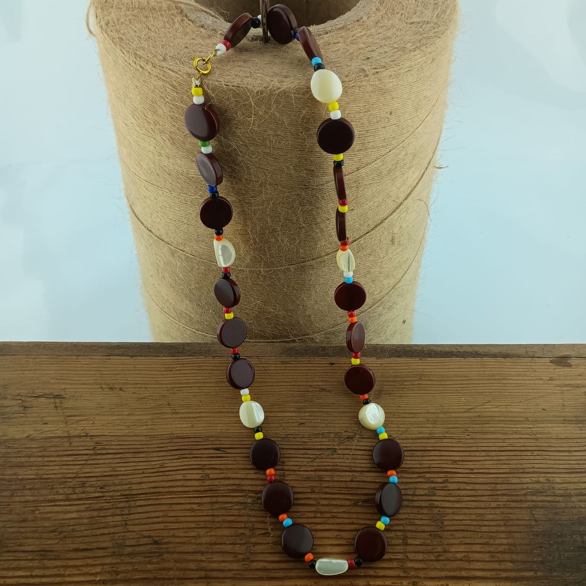 A beaded necklace with dark brown disc-shaped beads, white shell-like beads, and small colorful accent beads, displayed on a burlap-wrapped stand over a wooden surface.