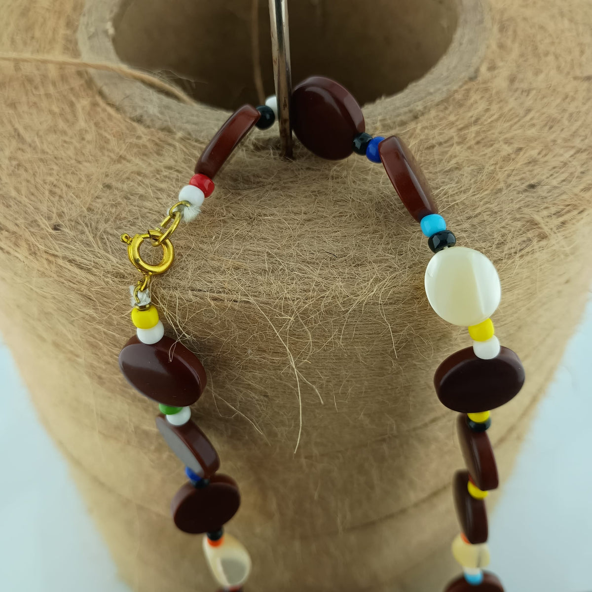A beaded necklace with dark brown polished oval beads, small multicolor spacer beads, and a gold clasp, displayed on a tan jute-covered jewelry stand.