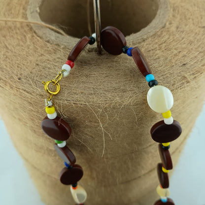 A beaded necklace with dark brown polished oval beads, small multicolor spacer beads, and a gold clasp, displayed on a tan jute-covered jewelry stand.
