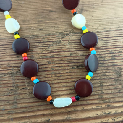 A beaded necklace with large dark reddish-brown round beads, white oval beads, and small multicolored spacer beads, displayed on a wooden surface.
